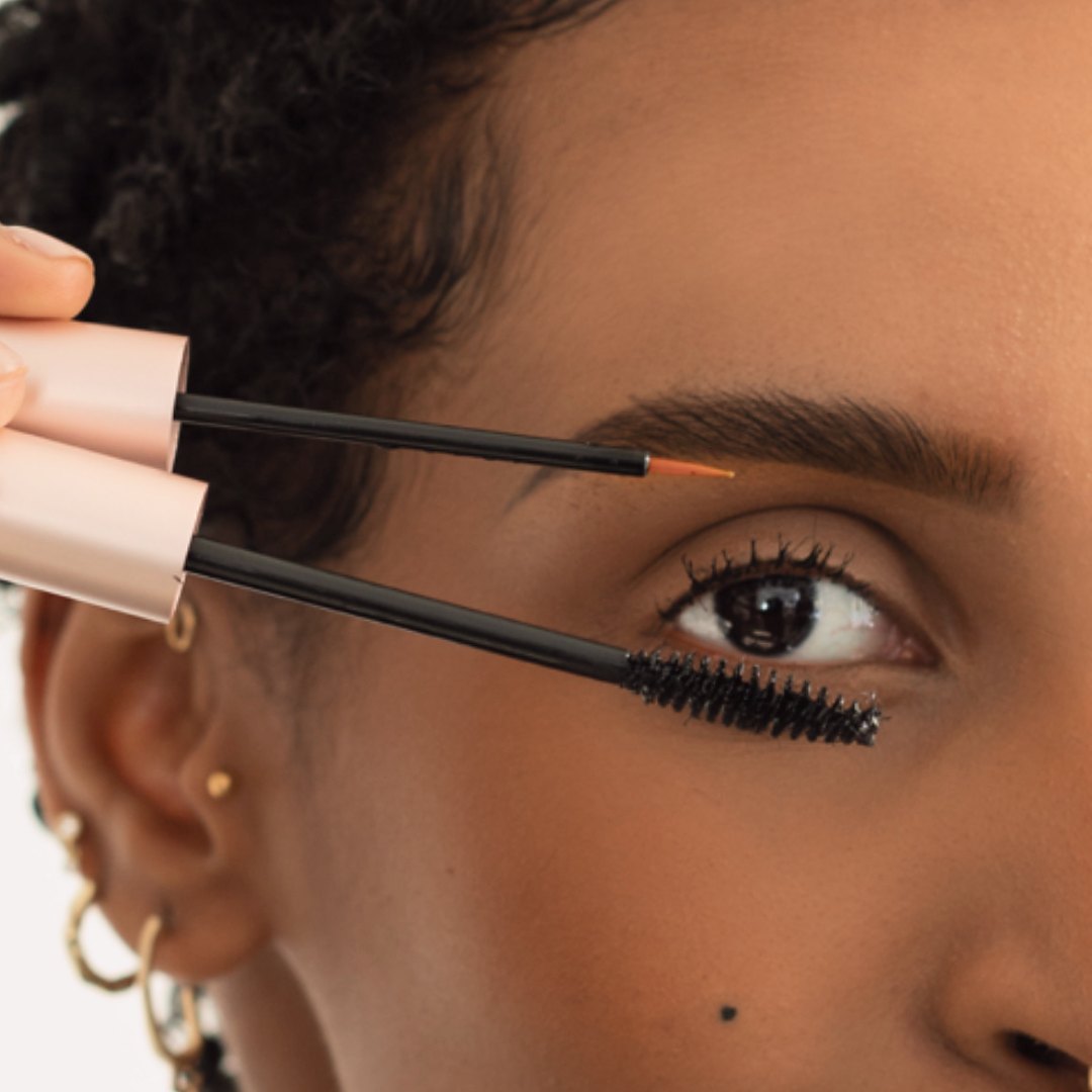 A woman holding the lash illusion serum and mascara wands at her lashes